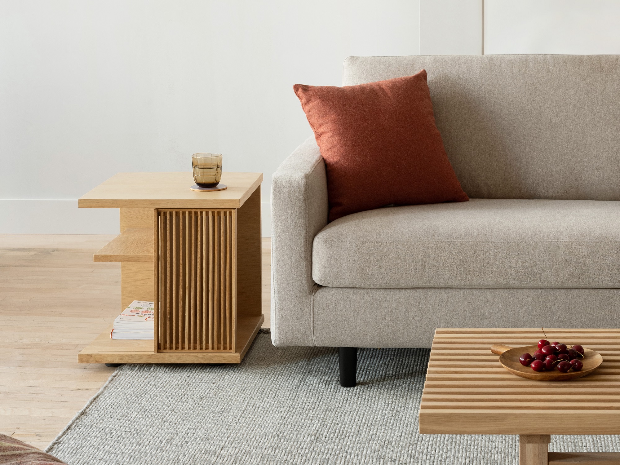 The Plank modern side table in oak with a glass on it sits beside a beige sofa and on a grey area rug